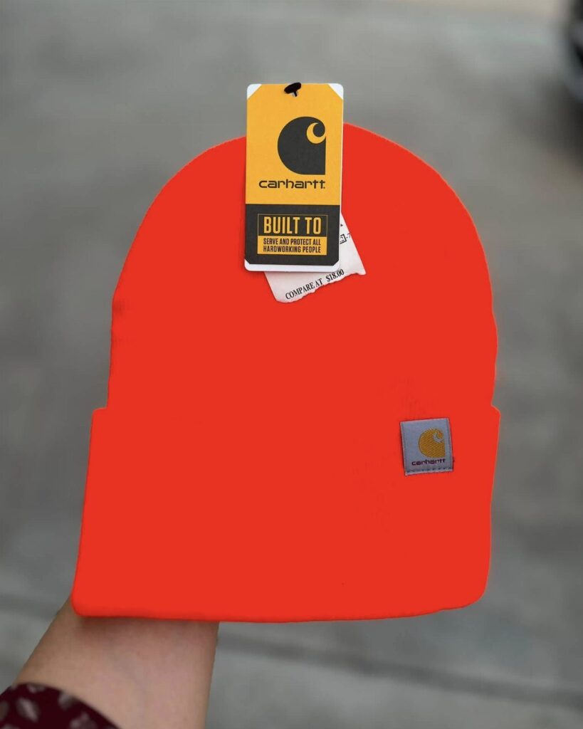 Orange Carhartt knit cuffed beanie with brand tag held against a grey background