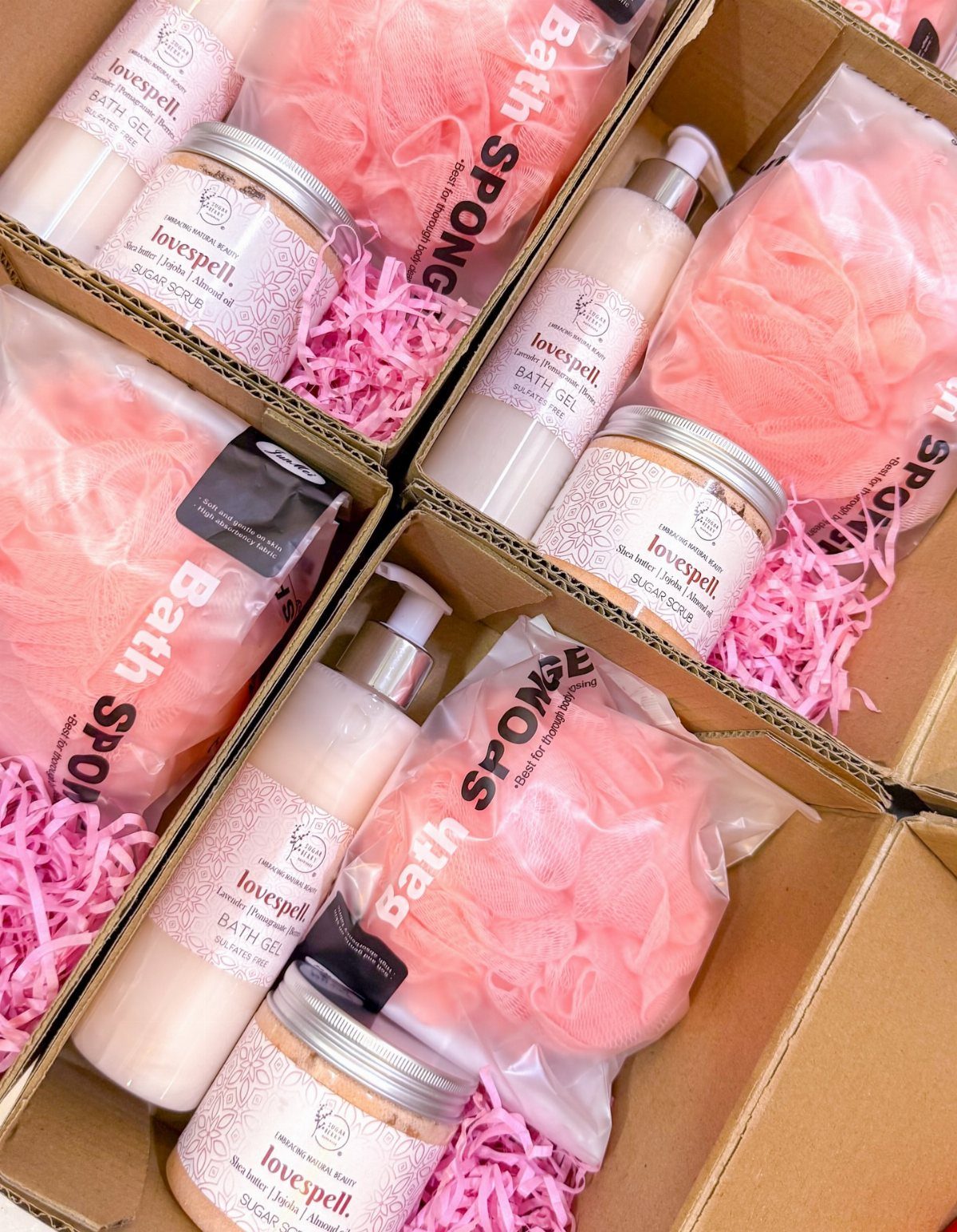 Four gift boxes containing pink bath sponges, sugar scrubs, and bath gel bottles.