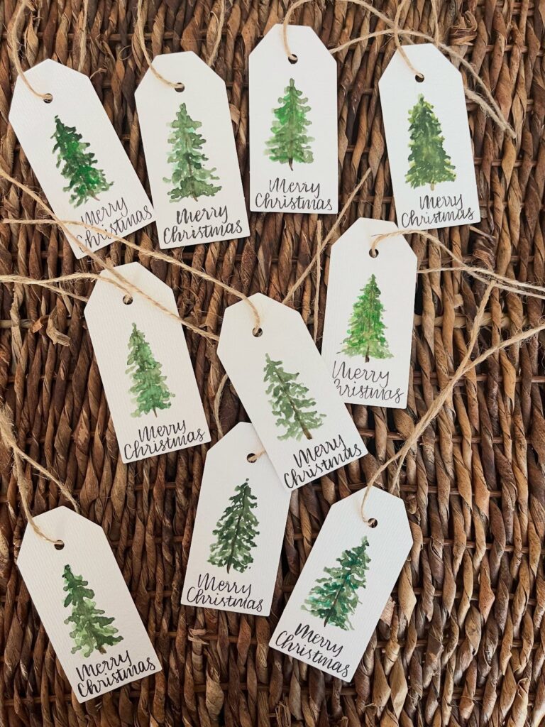 White Christmas gift tags with green watercolor trees strung on twine over wicker.