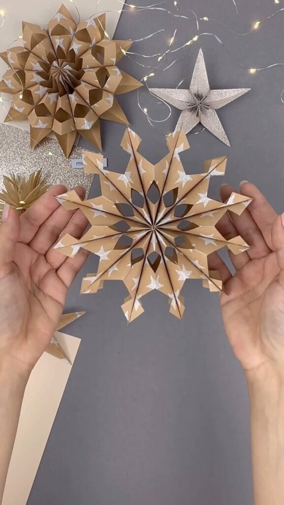 Hands holding a 3D folded brown paper snowflake with white star patterns.