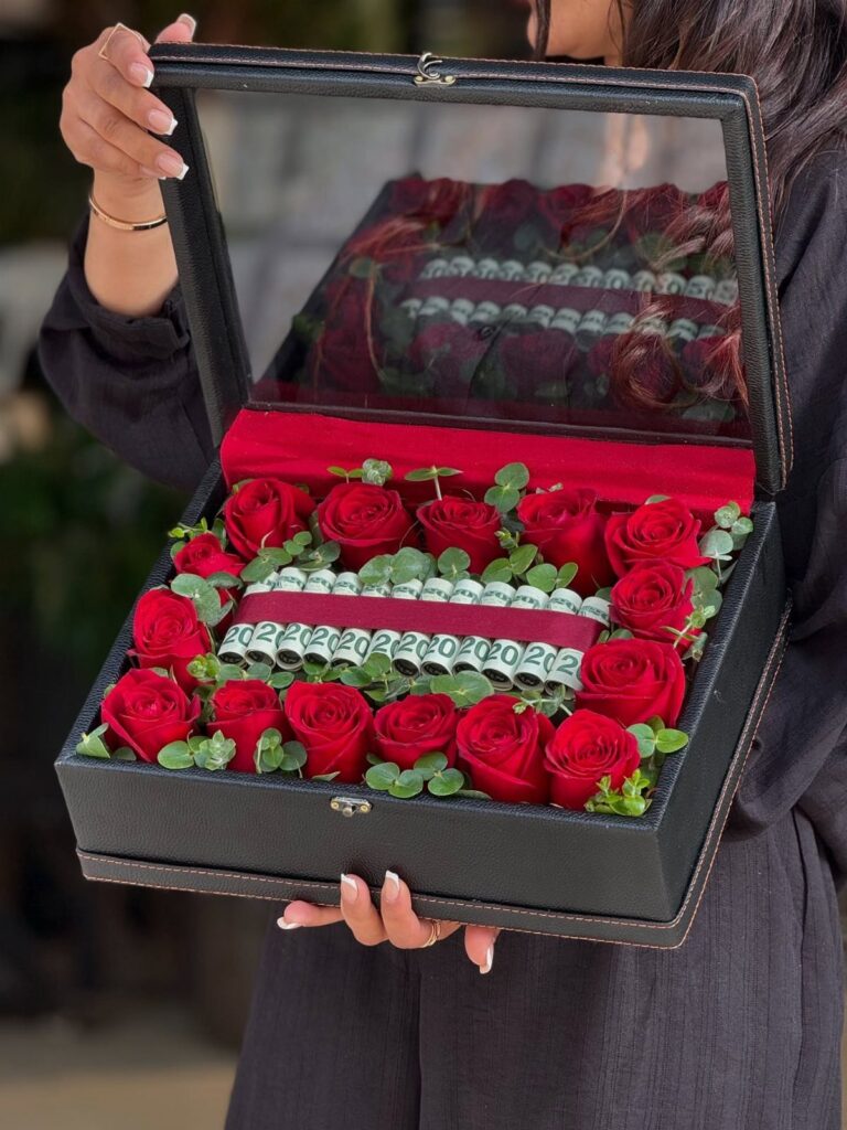 Black gift box filled with red roses and rolled-up cash banknotes.