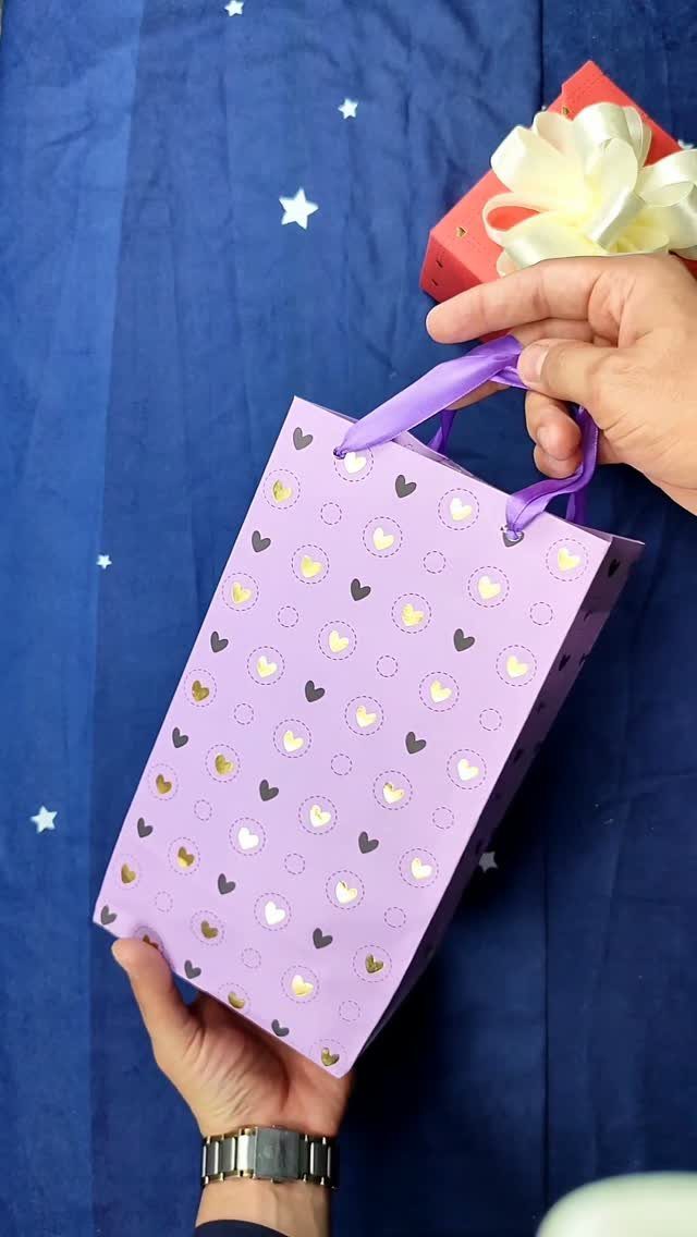 Purple heart-patterned gift bag with ribbon handles held by a person's hands.